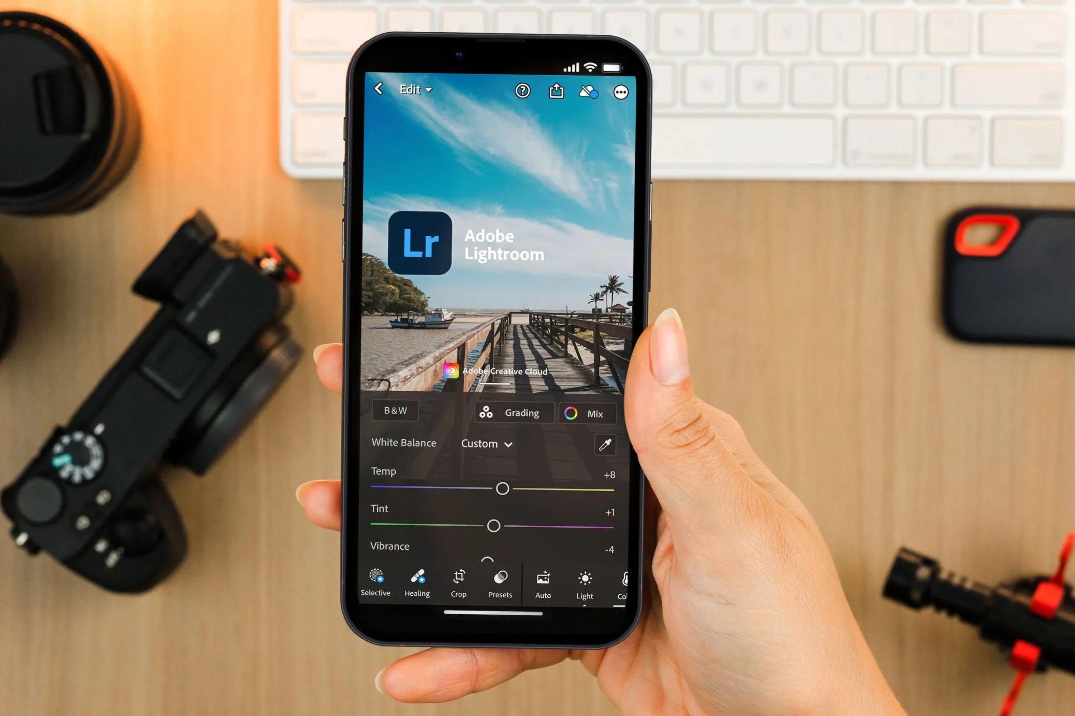 Bàn tay cầm điện thoại hiển thị ứng dụng Lightroom Mobile, phía sau là thiết bị máy ảnh, thể hiện tính năng chỉnh sửa ảnh chuyên nghiệp.